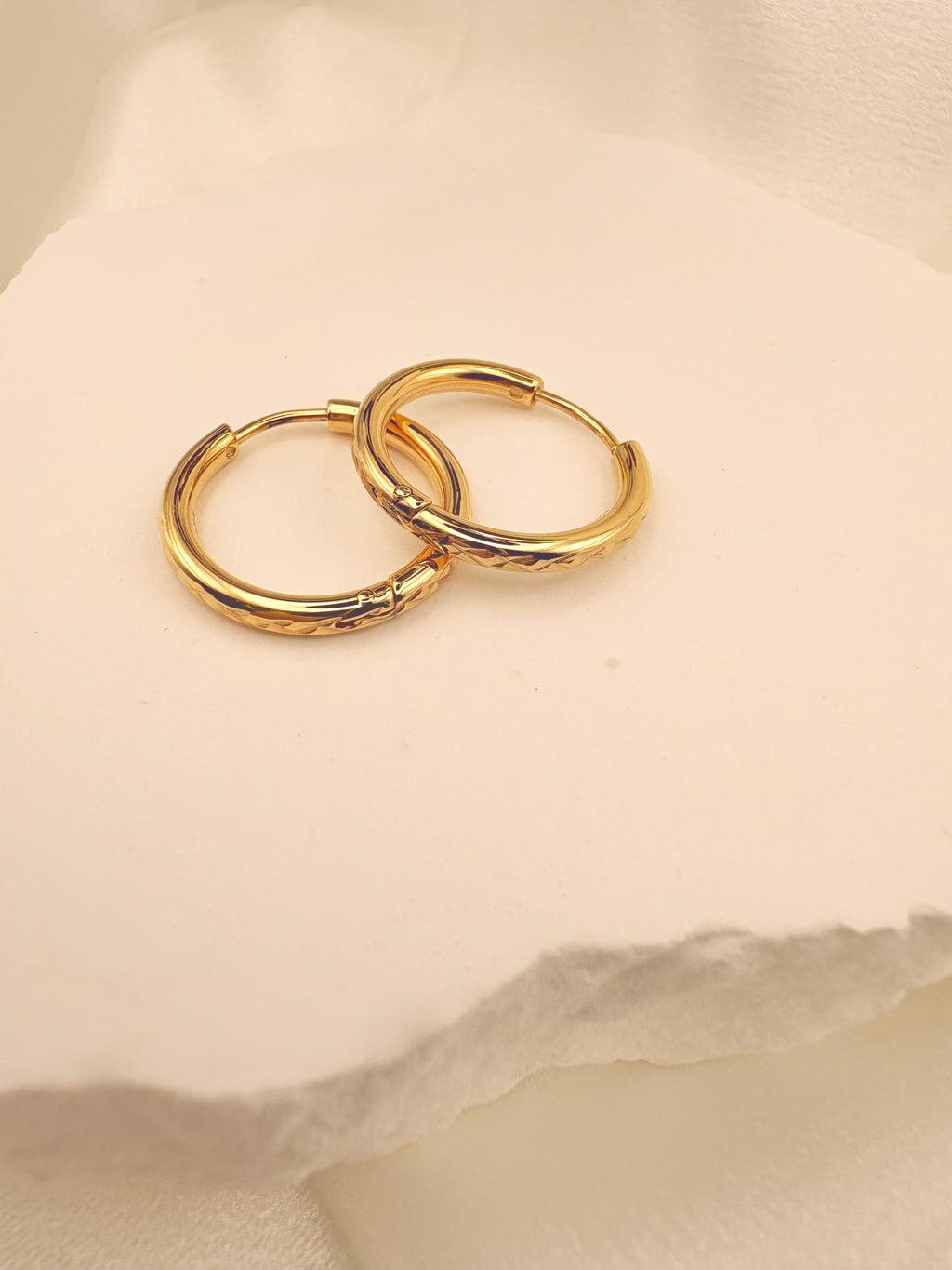 A pair of small gold hoop earrings with a textured, hammered finish, displayed on a soft, textured off-white surface.
