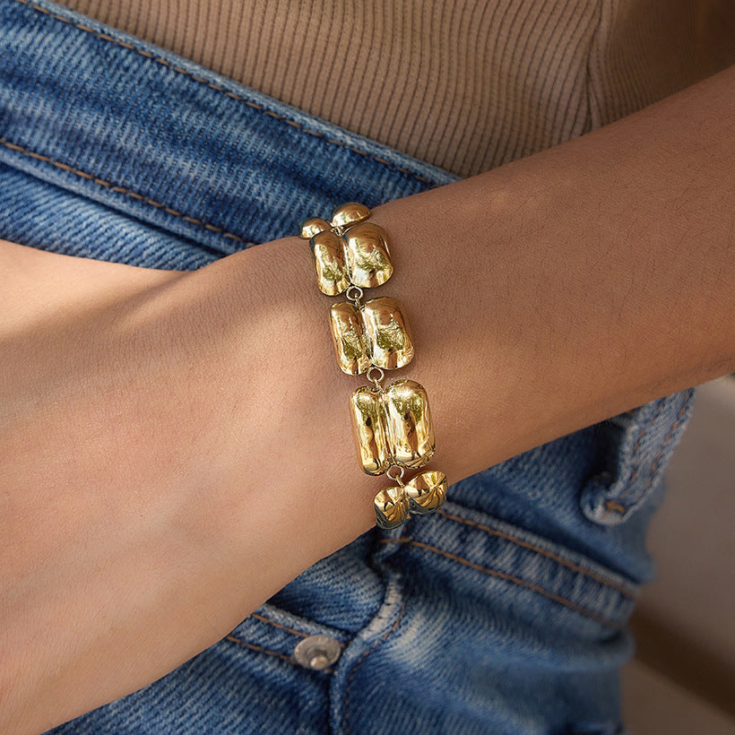 A close-up of a person's wrist wearing a chunky gold bracelet with large, irregular polished links, styled with a beige ribbed top and blue denim jeans.