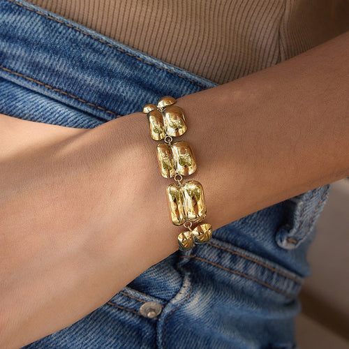 A close-up of a person's wrist wearing a chunky gold bracelet with large, irregular polished links, styled with a beige ribbed top and blue denim jeans.