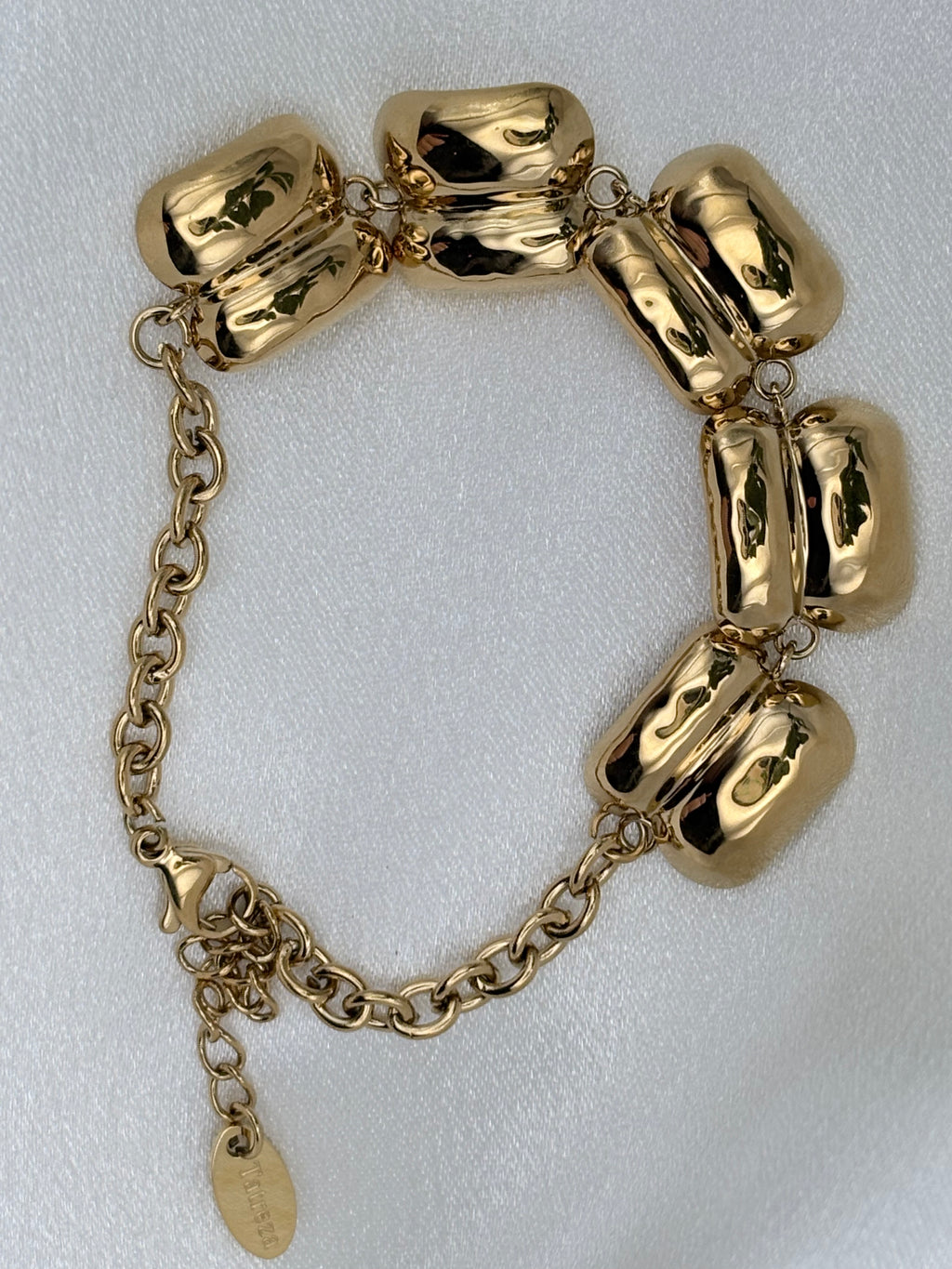 A gold-tone bracelet featuring large, polished, rounded links connected by a delicate chain, displayed on a textured white fabric background.
