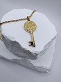 A gold key pendant necklace with an ornate circular keyhole design, resting on a textured white stone display against a light gray background.