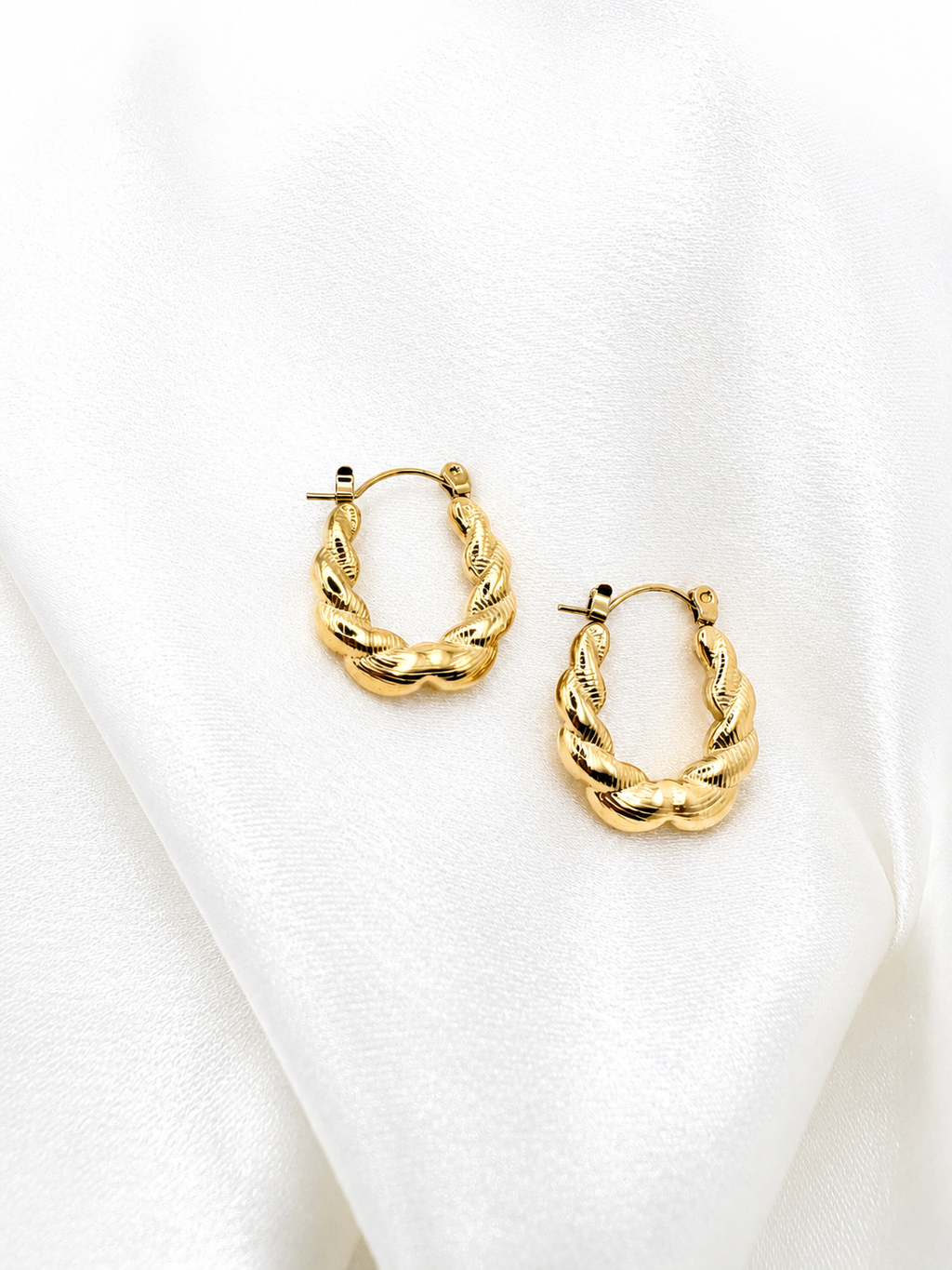 A pair of gold-toned twisted hoop earrings displayed on a draped white fabric background.
