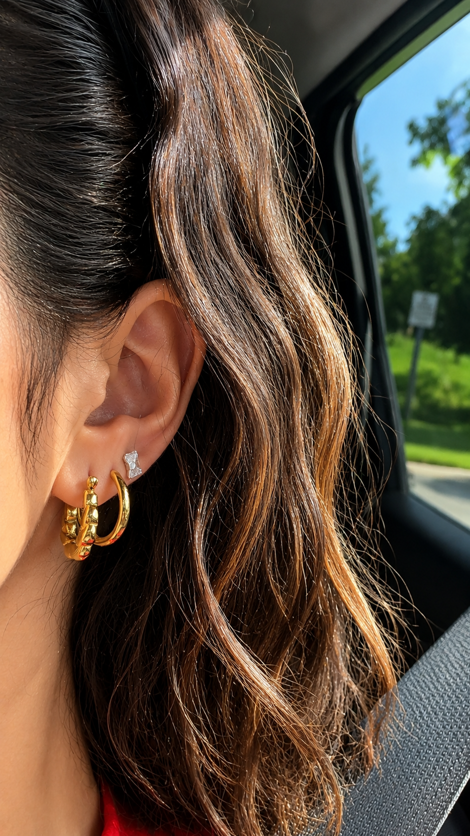 A close-up of a person wearing a pair of chunky gold hoop earrings with small clear stones, seated inside a car with the seatbelt visible.