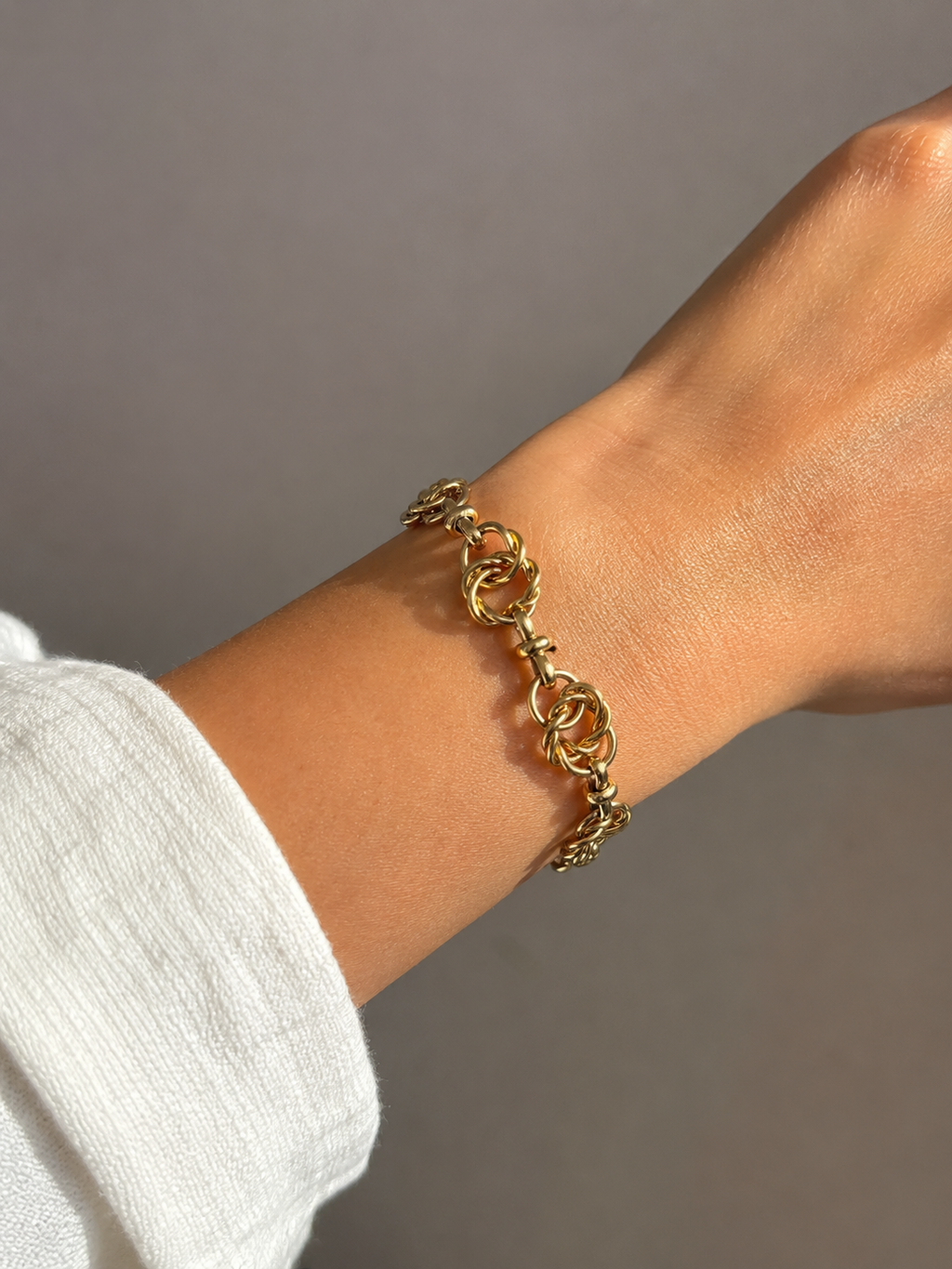 A gold chain link bracelet worn on a person's wrist, shown against a neutral gray background with a white sleeve visible.