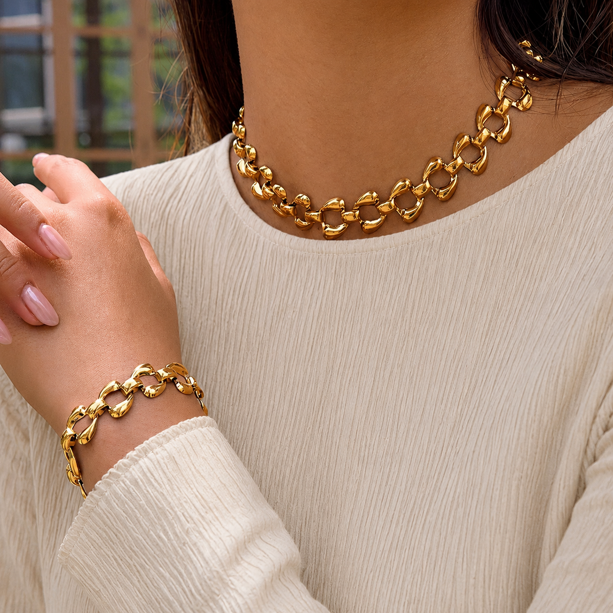 A close-up of a person wearing a chunky gold chain necklace and a matching gold chain bracelet, paired with a cream-colored ribbed top.