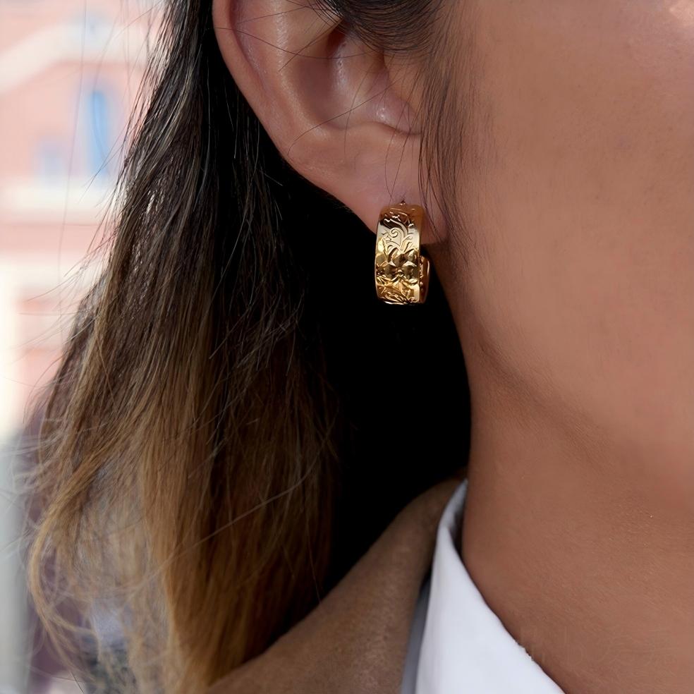 A close-up of a person's ear wearing a textured gold huggie hoop earring, with dark hair and a white collared shirt visible against a softly blurred background.