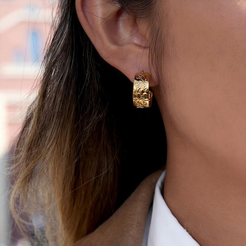 A close-up of a person's ear wearing a textured gold huggie hoop earring, with dark hair and a white collared shirt visible against a softly blurred background.